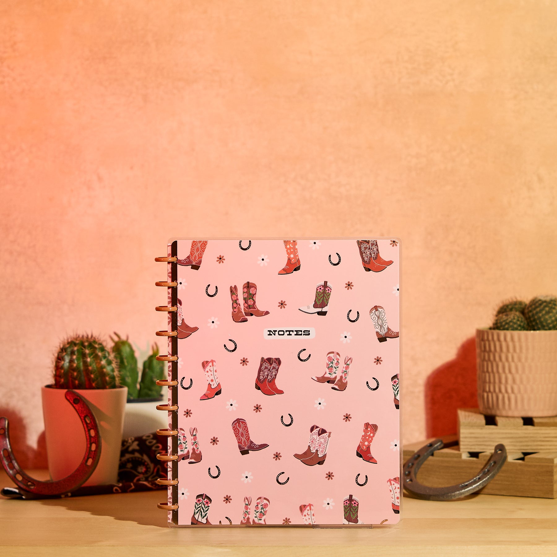 Western Bloom - Dotted Lined Big Notebook - 60 Sheets with pink cover featuring colorful cowboy boots and horseshoe designs, standing upright on a wooden surface with decorative cacti and horseshoes in the background.
