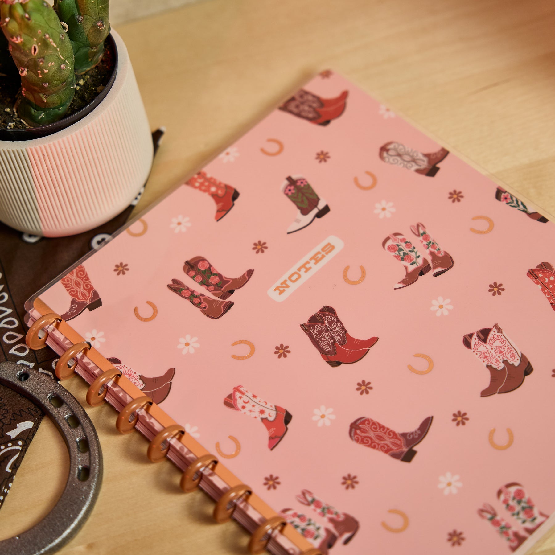 Western Bloom - Dotted Lined Big Notebook - 60 Sheets with a pink cover featuring colorful western boot patterns, a flexible disc-bound spine, placed on a wooden surface next to a potted cactus and decorative horseshoe.