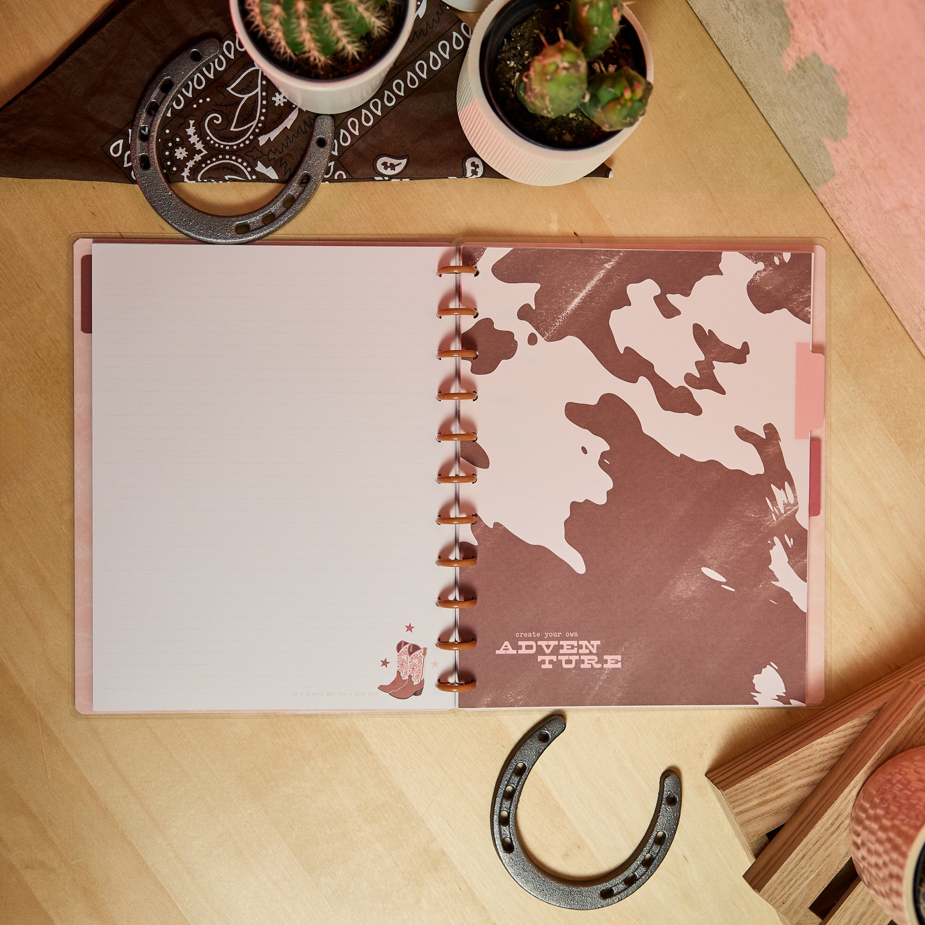 Open Western Bloom - Dotted Lined Big Notebook - 60 Sheets lying on a wooden table next to potted cacti, horseshoes, and a brown bandana, featuring a western cowboy boot design on the lined pages.