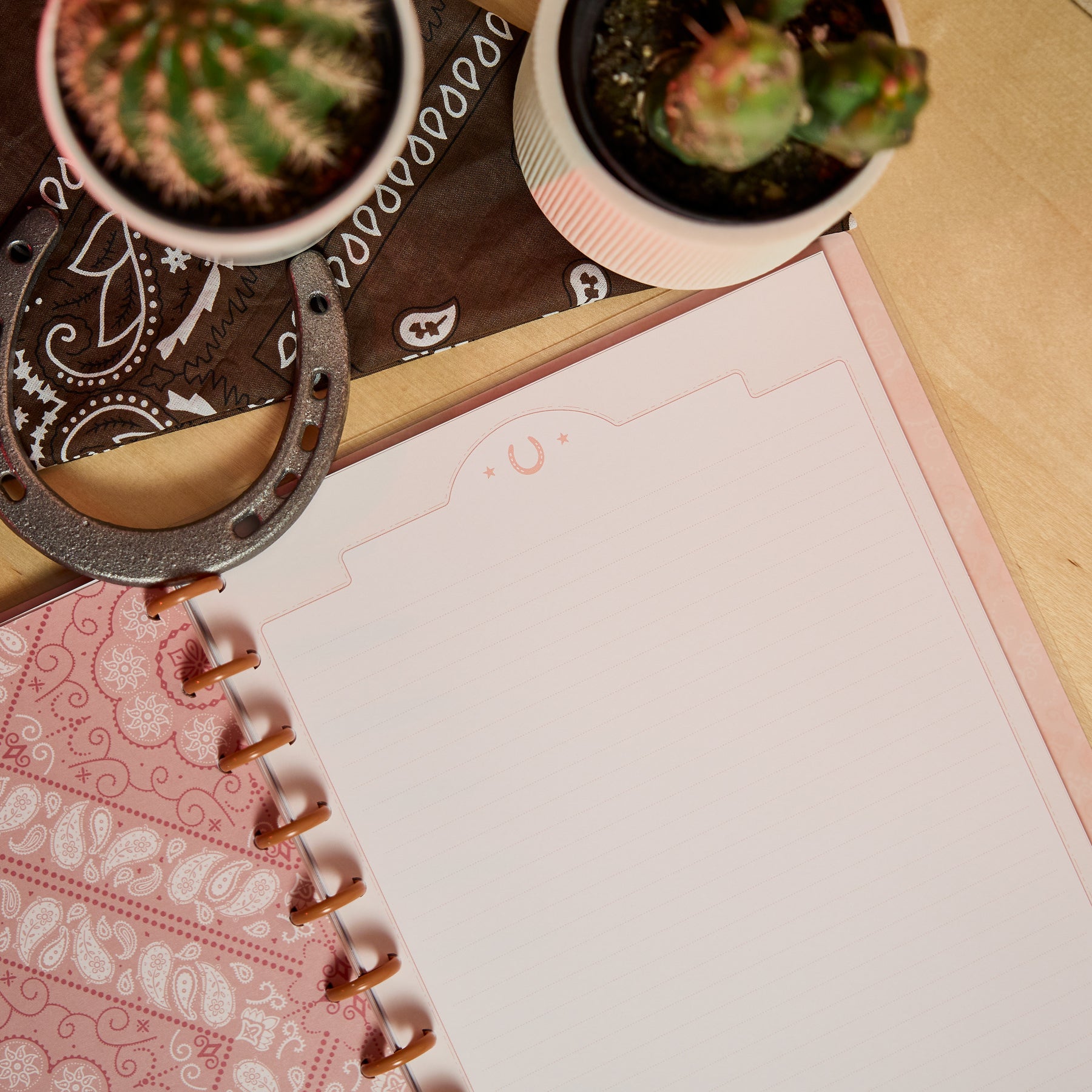 Open Western Bloom - Dotted Lined Big Notebook - 60 Sheets with pink disc binding on a wooden surface, next to small potted cacti and a horseshoe on a brown patterned cloth.