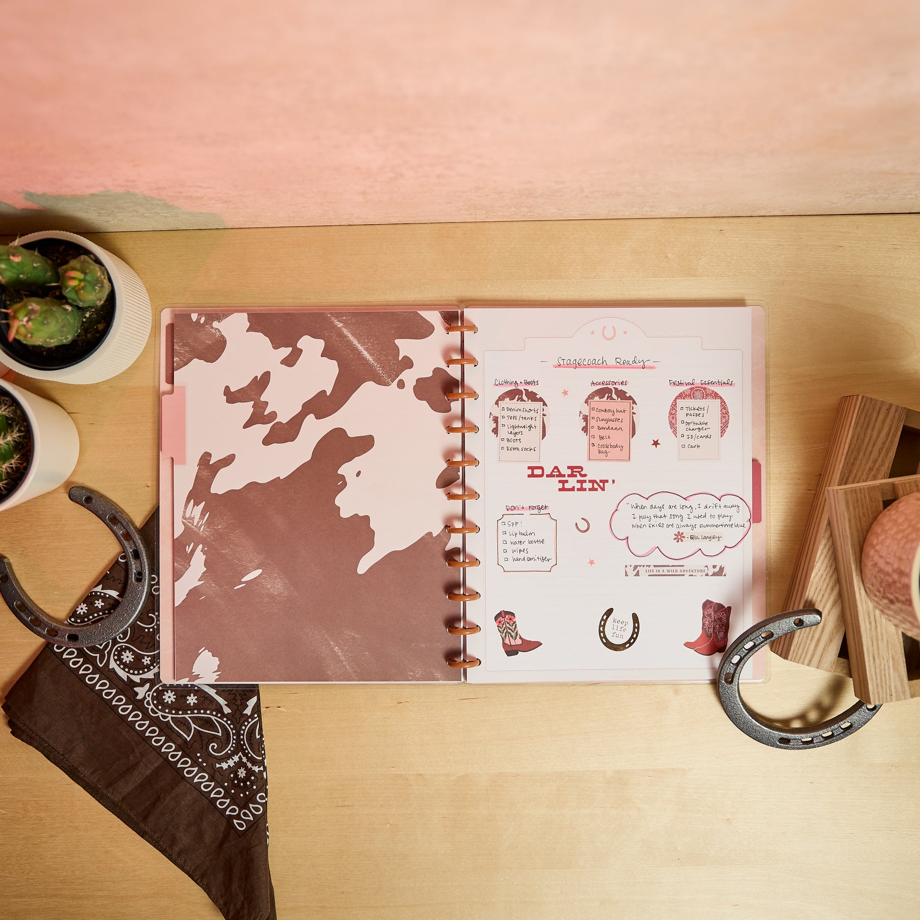 Open Western Bloom - Dotted Lined Big Notebook - 60 Sheets showing a cowhide pattern cover with rose gold discs, styled with cowboy boots, horseshoes, and cacti on a wooden surface.