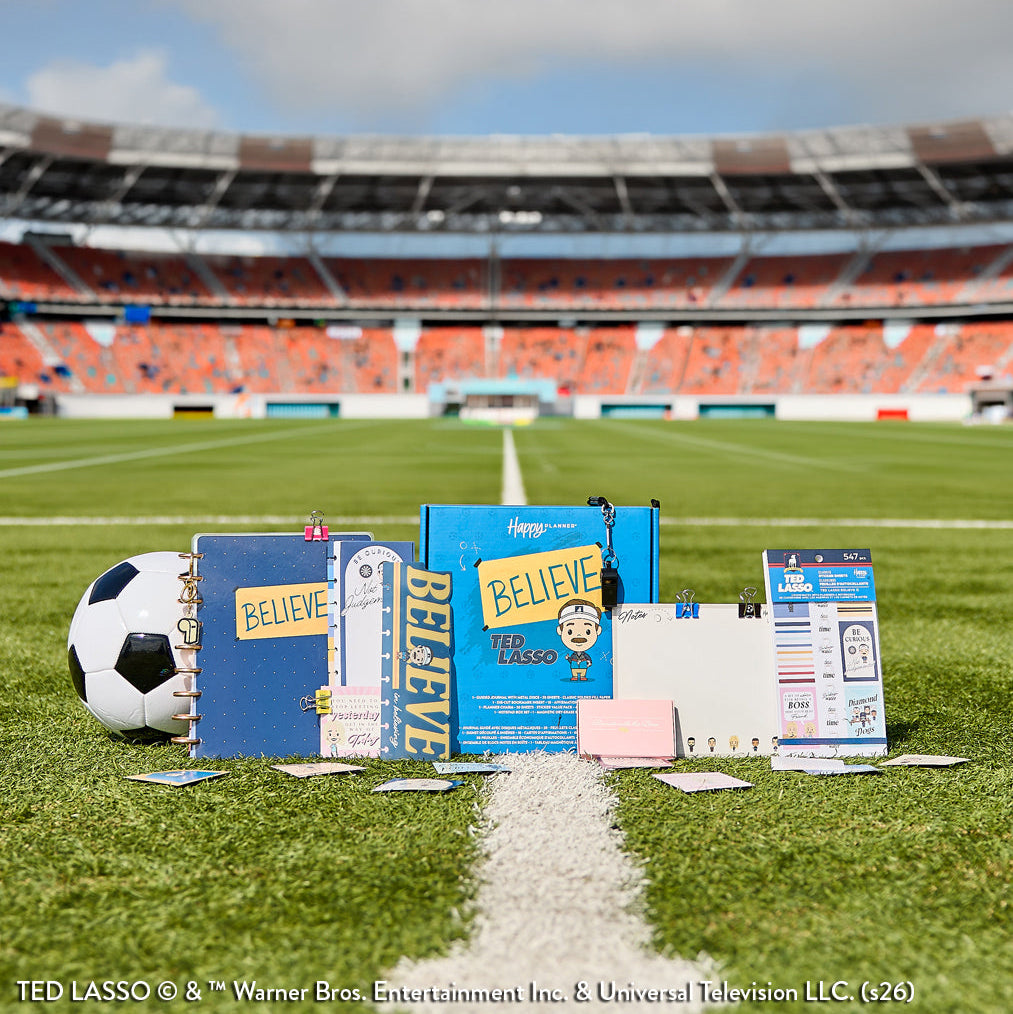 Ted Lasso - Be Happy Box displayed on a soccer field with a soccer ball, featuring disc-bound planners, guided journals, stickers, and happy planner accessories in shades of blue and yellow.