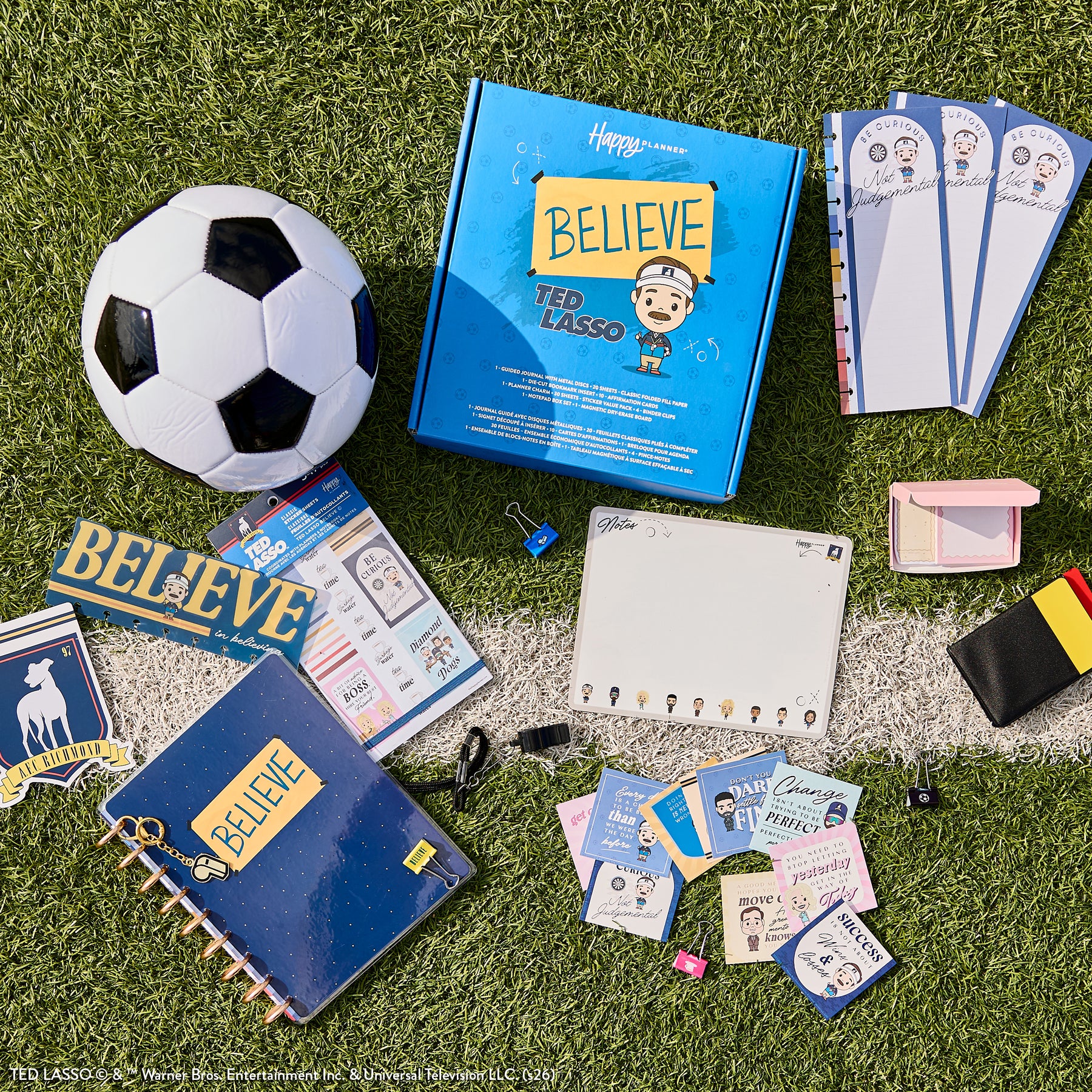 Flat lay of Ted Lasso - Be Happy Box featuring a blue Happy Planner binder with "BELIEVE" text, soccer ball, themed stickers, notebooks, bookmarks, and planner accessories on grass and concrete surface.