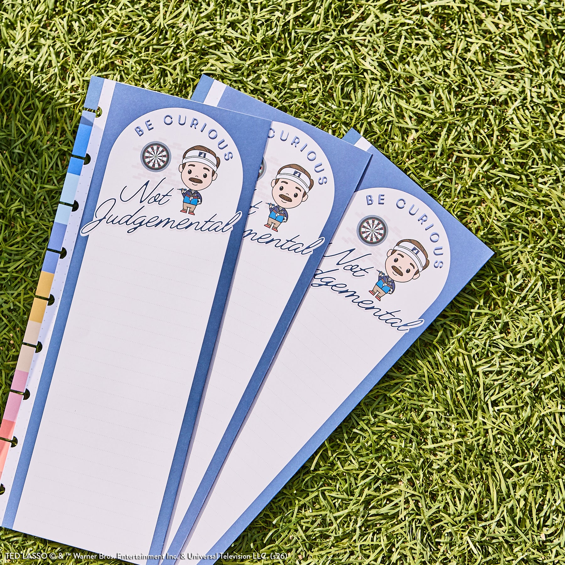Three blue pages with a cartoon character and the text "Be Curious Not Judgemental" lie on grass, designed as part of the Ted Lasso - Be Happy Box planner set.