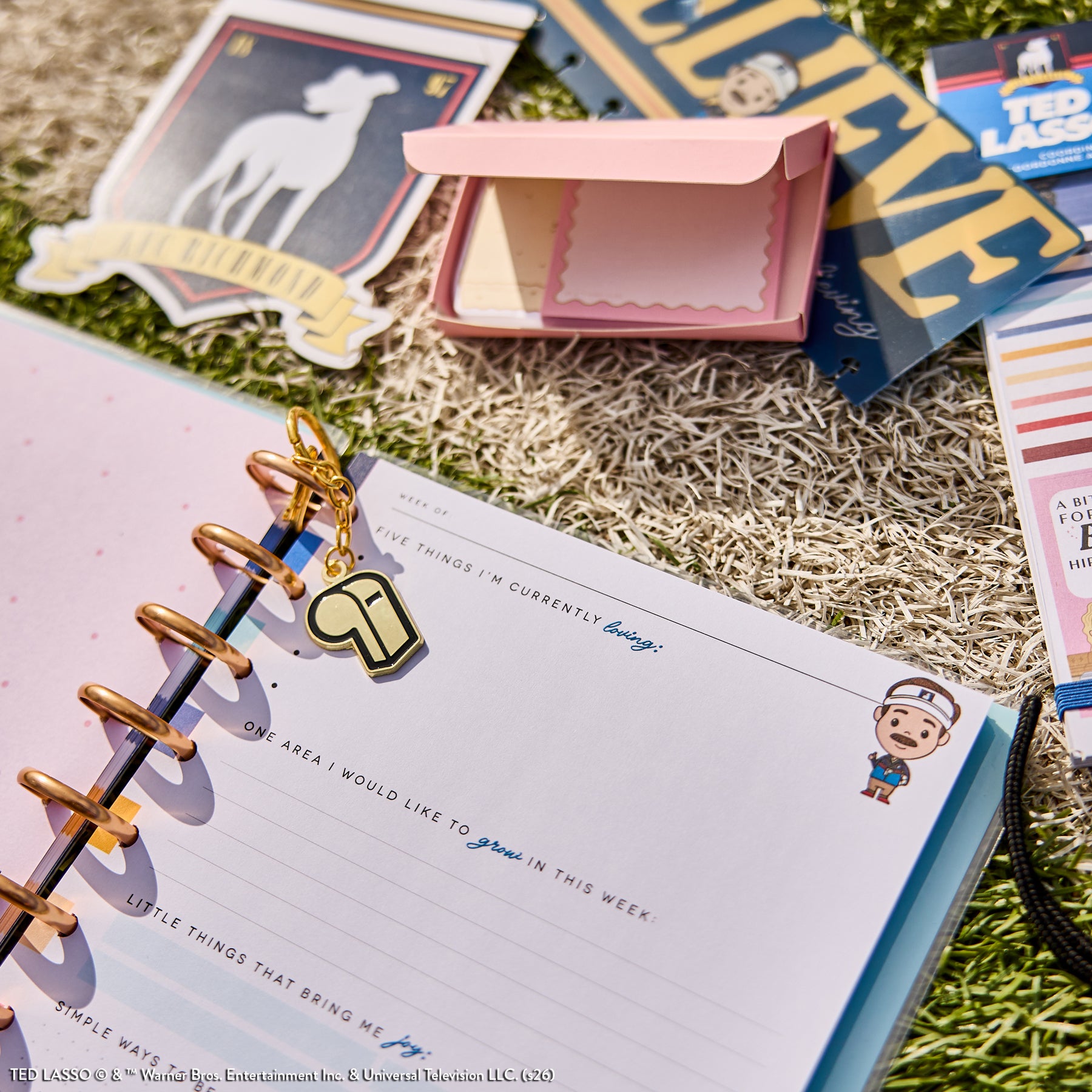 Open Ted Lasso - Be Happy Box planner with disc bound pages, featuring prompts for gratitude and goals, surrounded by stickers and related paper goods on grass outdoors.