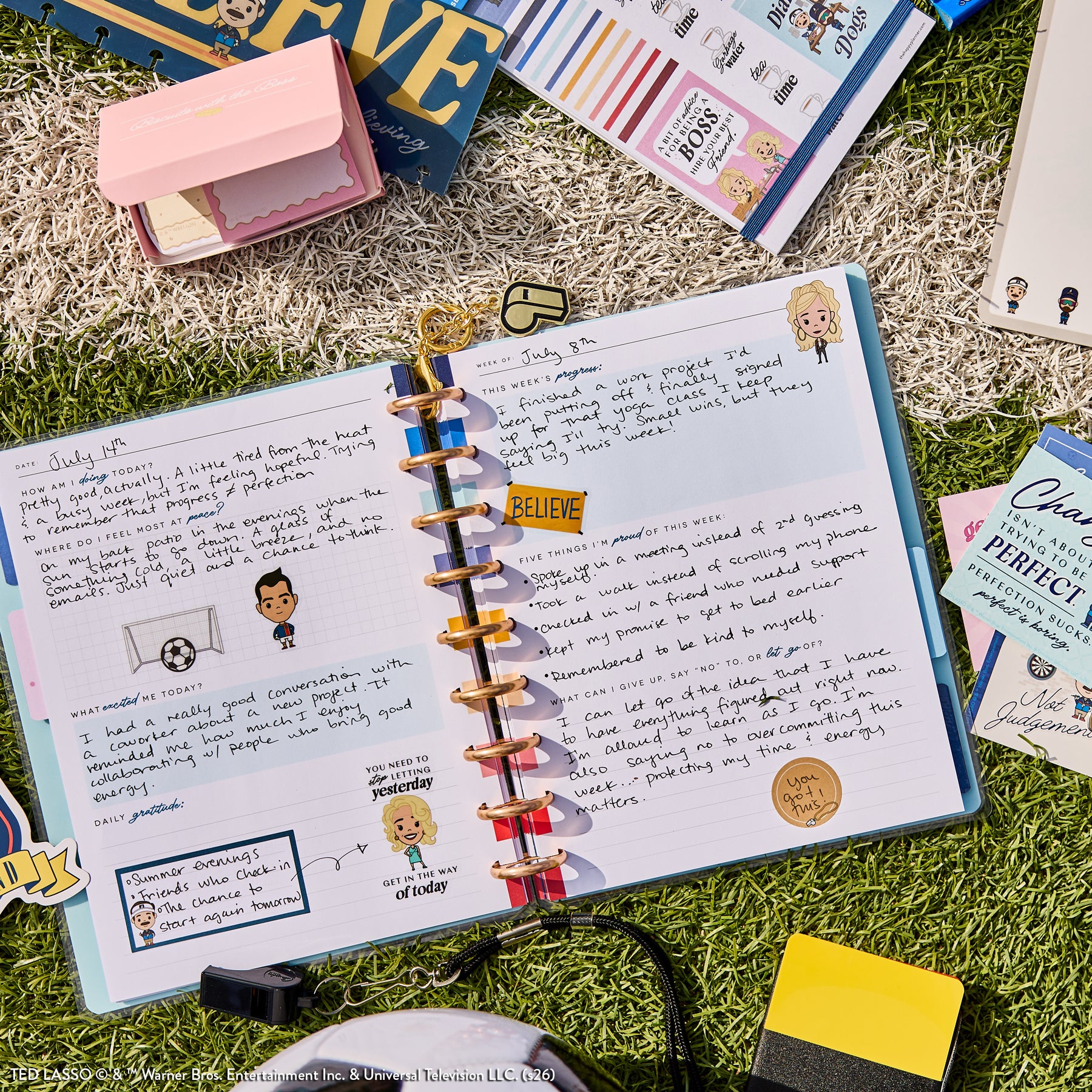 Open disc-bound planner spread with handwritten notes, stickers, and colorful tabs, featuring characters and motivational elements from Ted Lasso - Be Happy Box on grass background.