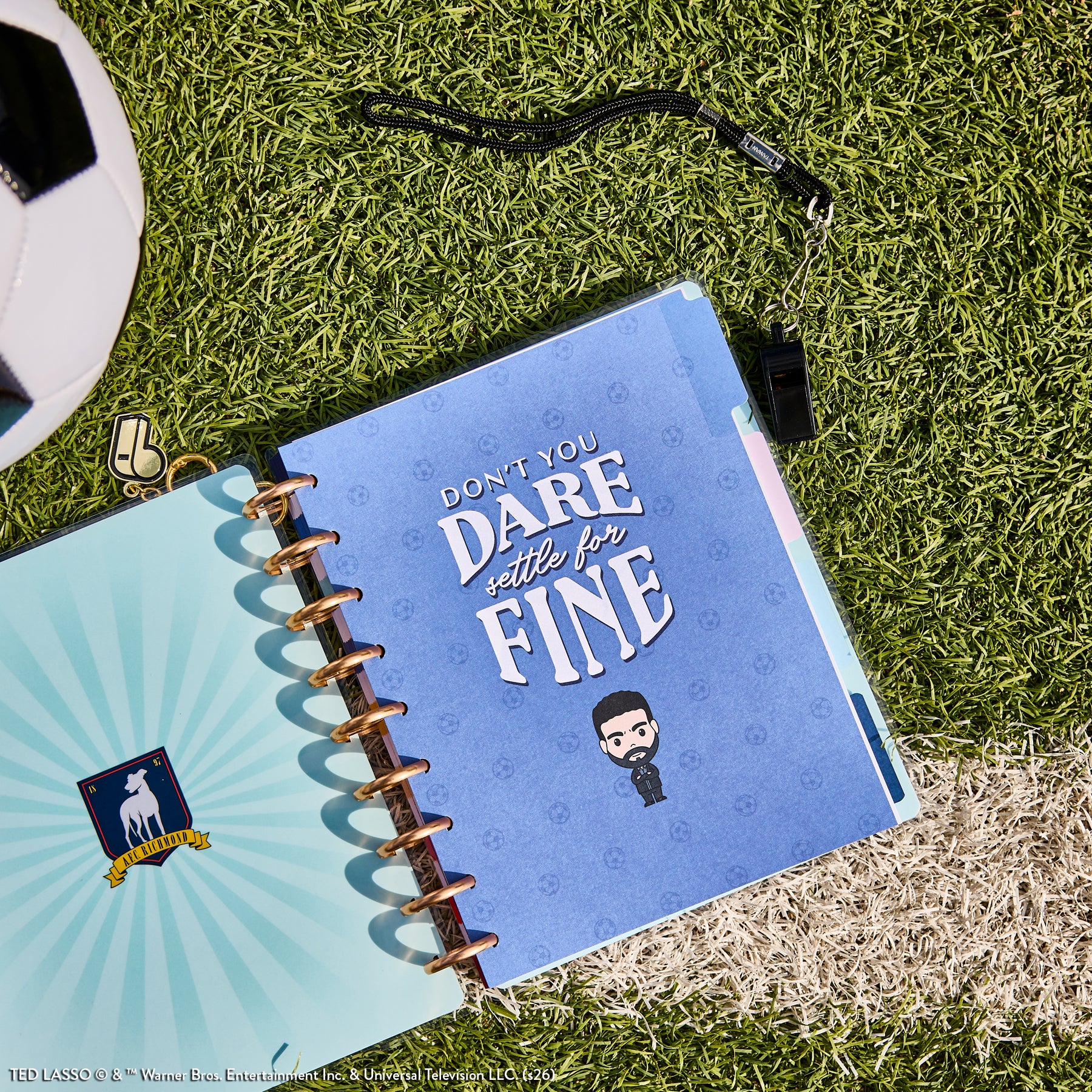Ted Lasso - Be Happy Box disc-bound planner open on artificial grass next to a soccer ball, featuring a blue cover with the quote "Don&