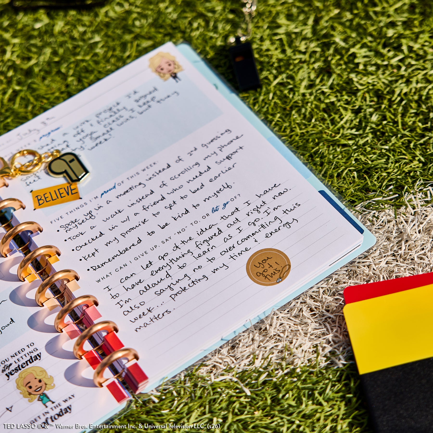 Open disc-bound planner from the Ted Lasso - Be Happy Box showing handwritten notes, stickers, and a whistle keychain on a grass-like surface with colorful card sleeves nearby.