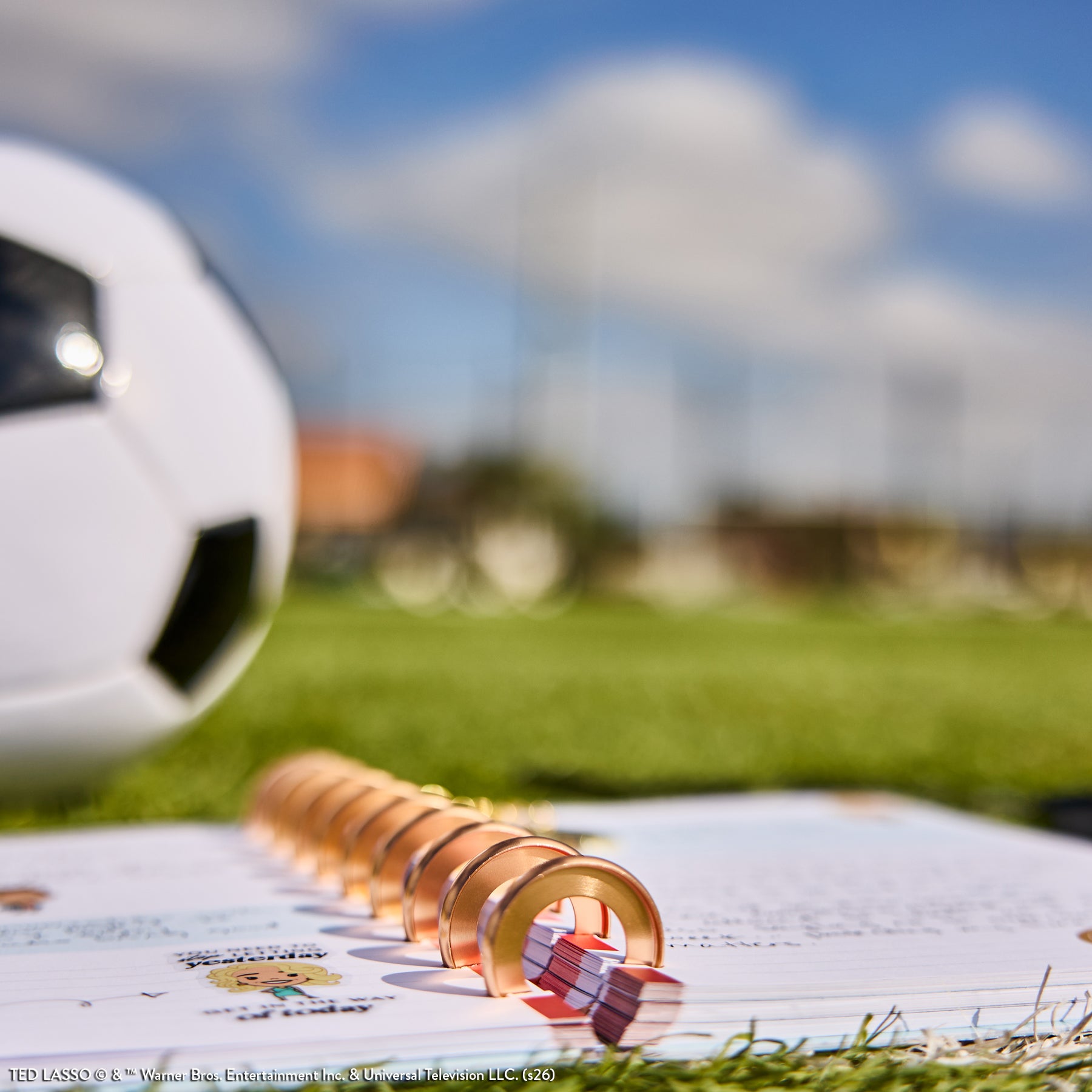 Close-up of an open disc-bound planner with rose gold discs lying on grass near a soccer ball under a blue sky, featuring the Ted Lasso - Be Happy Box collection.