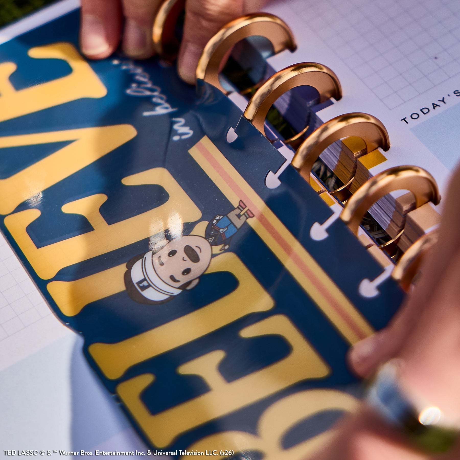 Close-up of a hand inserting a clear plastic page featuring a Ted Lasso character into a gold disc-bound planner from the Ted Lasso - Be Happy Box collection.