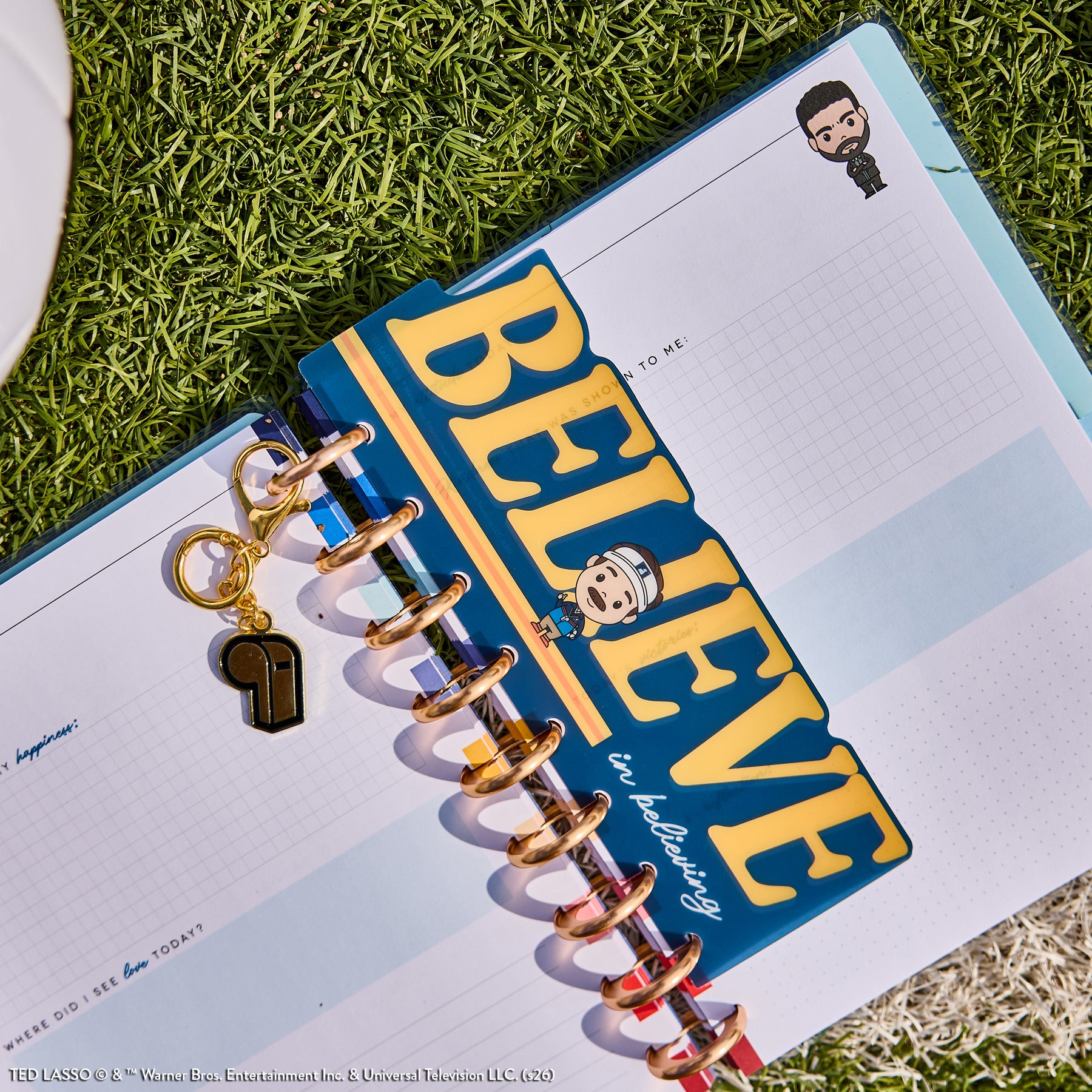 Ted Lasso - Be Happy Box open disc-bound planner showing a colorful "BELIEVE" bookmark with cartoon characters and a gold whistle charm on the discs, resting on grass.