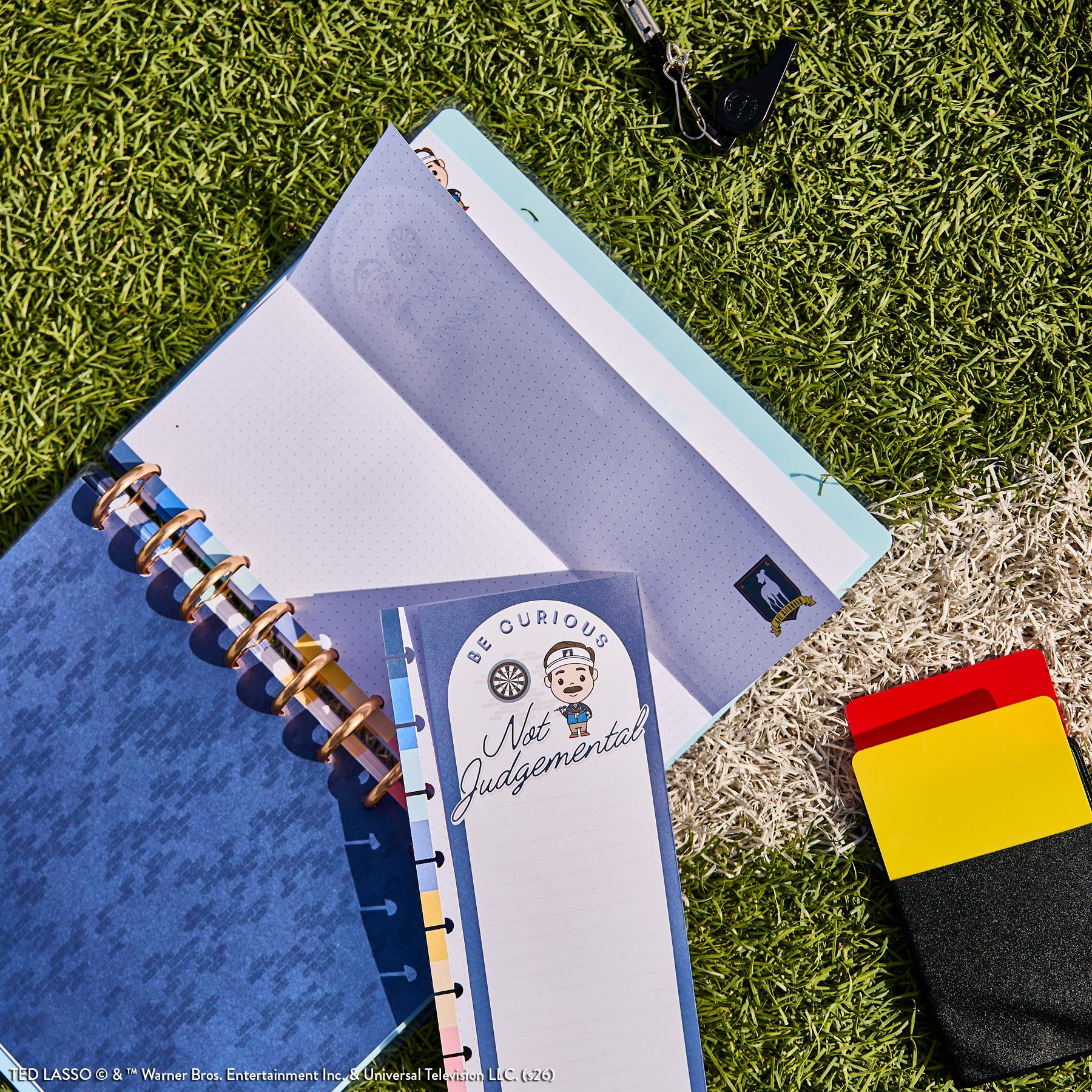 Ted Lasso - Be Happy Box open disc-bound planner with guided journal pages featuring motivational phrases, lying on grass next to a pair of black and yellow athletic socks.
