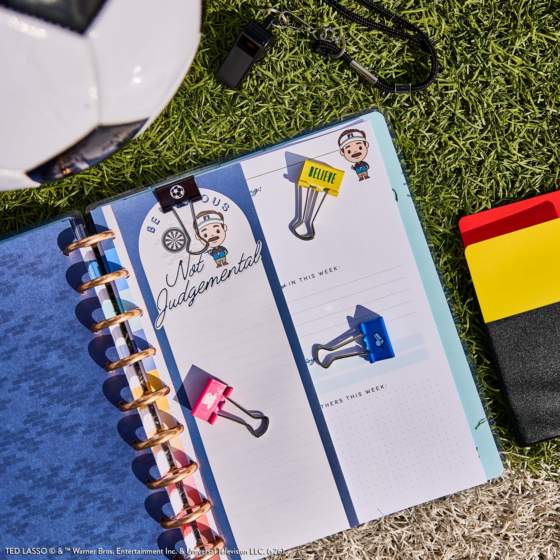 Open Ted Lasso - Be Happy Box planner on grass with colorful binder clips, a whistle, and a soccer ball nearby, showcasing happy planner pages with motivational details.