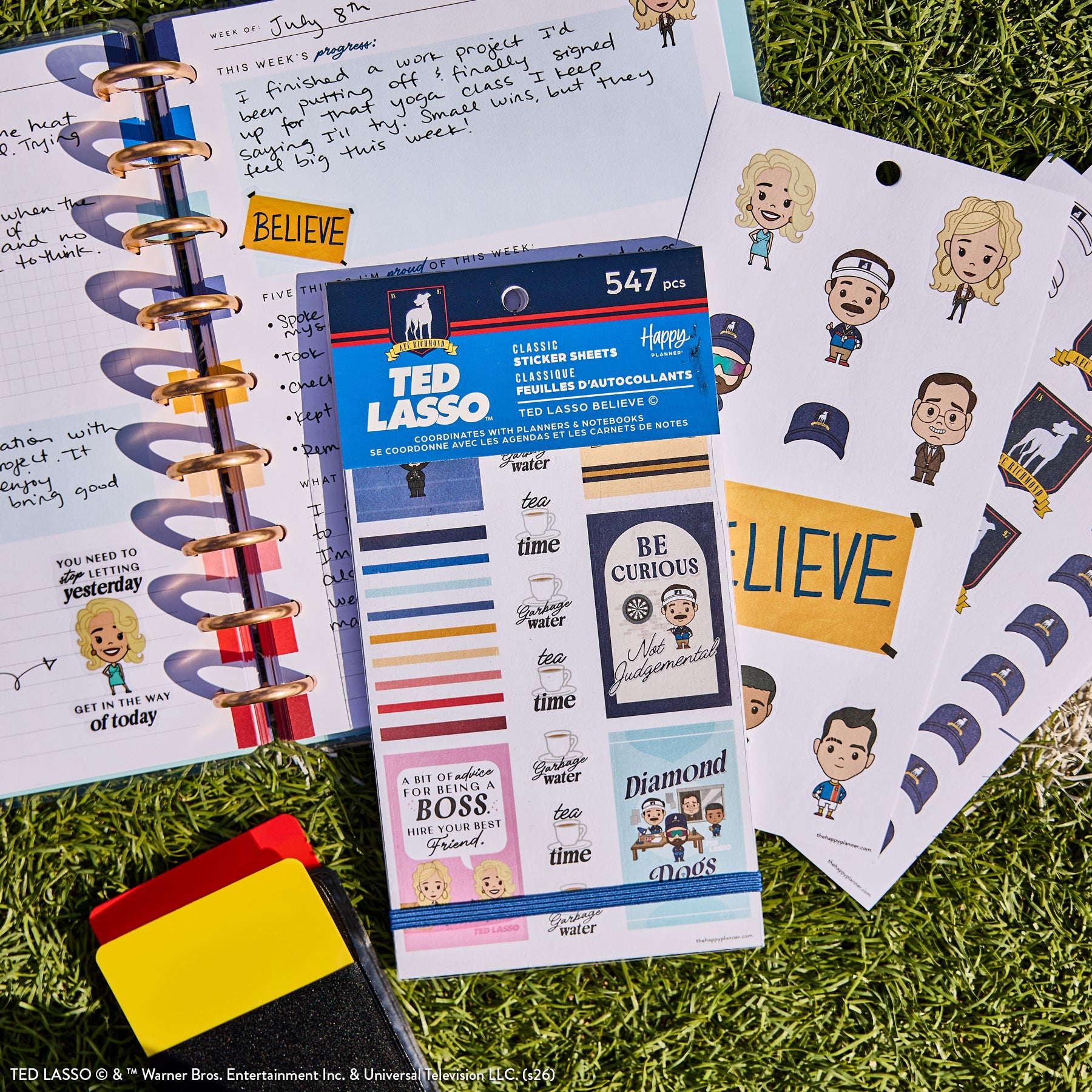 Ted Lasso - Be Happy Box sticker sheets featuring characters, motivational phrases, and themed designs displayed on a planner with colorful disc binding on grass background.