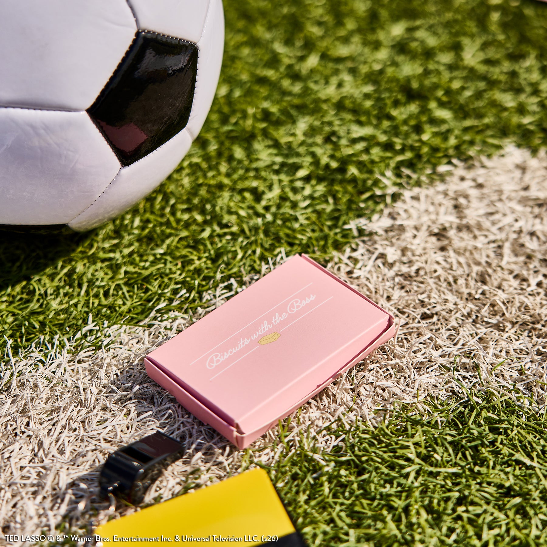 A soccer ball on grass next to a pink box with the text "Ted Lasso - Be Happy Box" placed near a black clip and a yellow planner on the edge of a white field line.