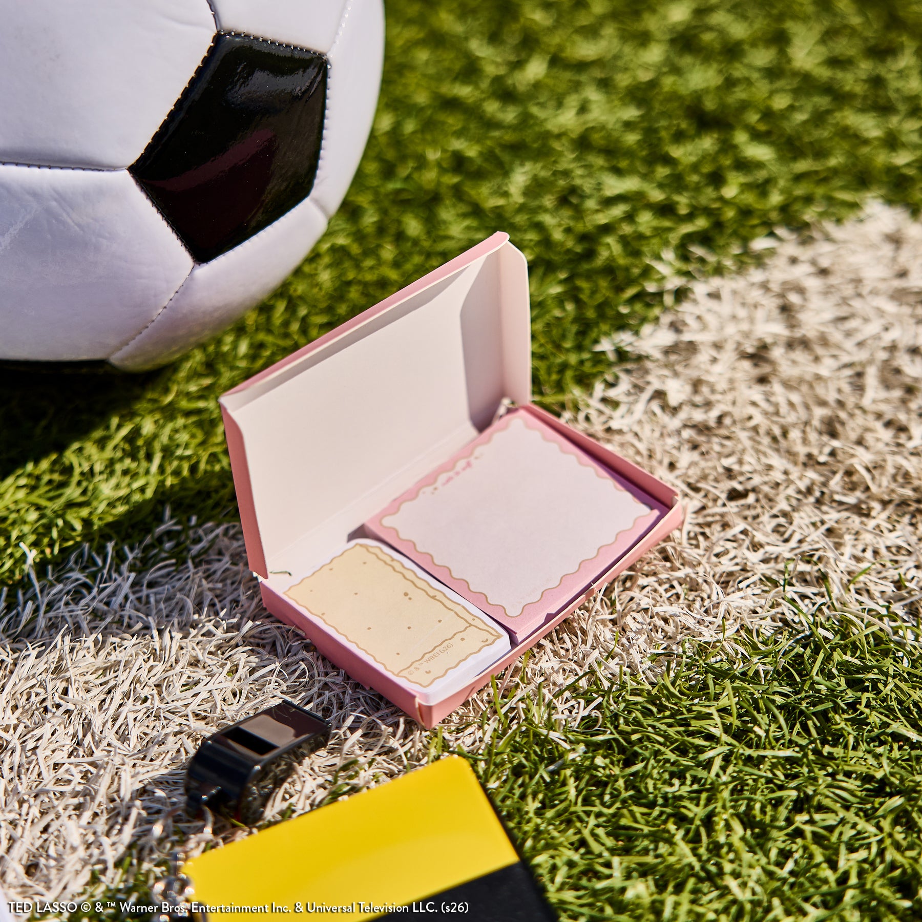 Ted Lasso - Be Happy Box featuring a pink open box with sticky notes and a black whistle next to a soccer ball on grass, evoking sports and planning themes.