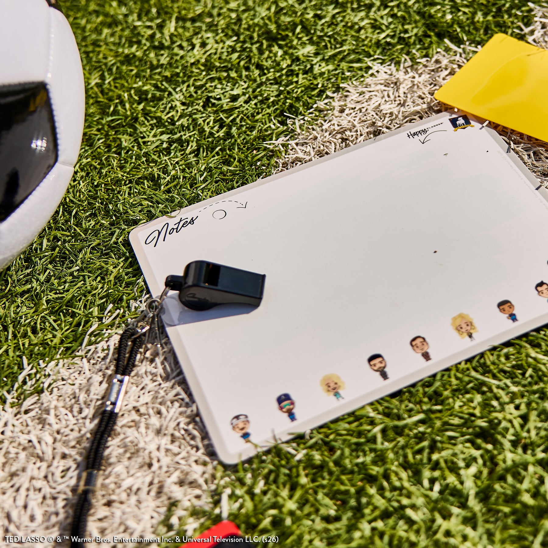 Whiteboard note pad with Ted Lasso character illustrations, a black whistle, and a yellow card on artificial grass near a soccer ball, featuring Ted Lasso - Be Happy Box.