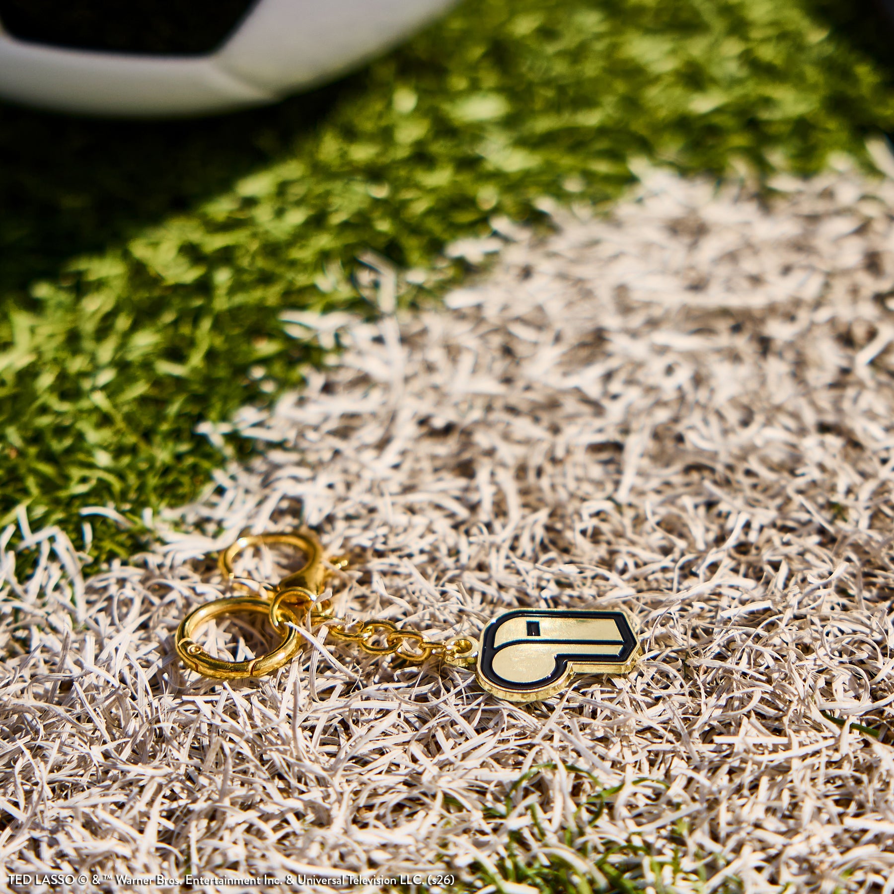Close-up of a gold whistle keychain charm on textured grass, part of the Ted Lasso - Be Happy Box collection featuring small accessories inspired by the series.